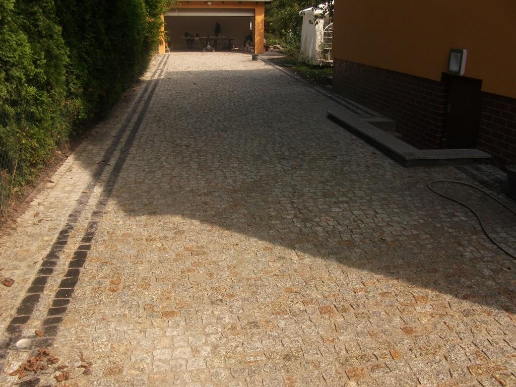 Ułożona kostka granitowa w odcieniach beżu i brązu tworzy ścieżkę w ogrodzie, z ciemnymi pasami po bokach. W tle widoczny budynek z żółtą elewacją i garaż.