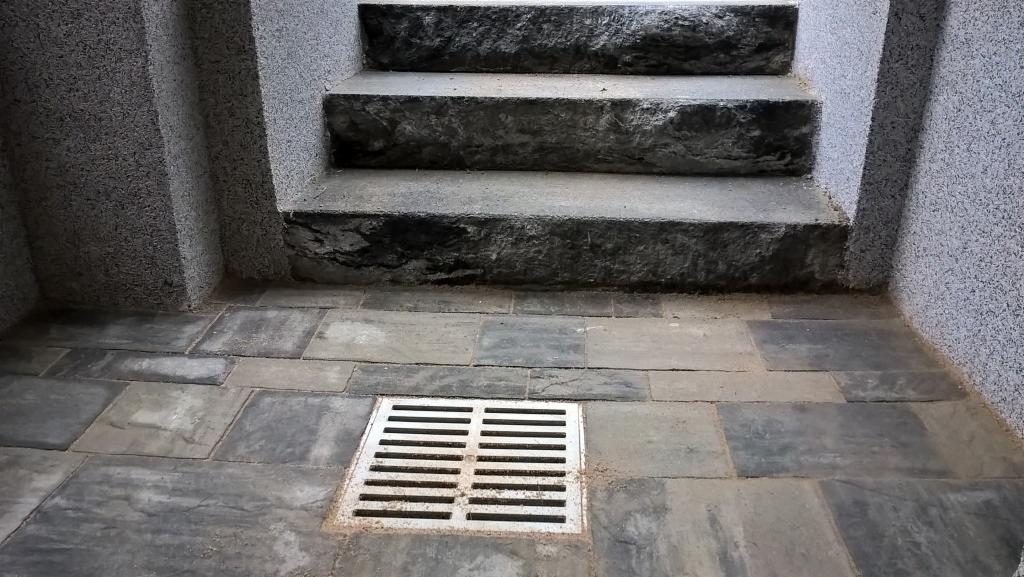 Kamienne schody z odpływem liniowym i szarą kostką brukową tworzą eleganckie przejście. Teksturowane ściany dodają charakteru. Detal architektoniczny.