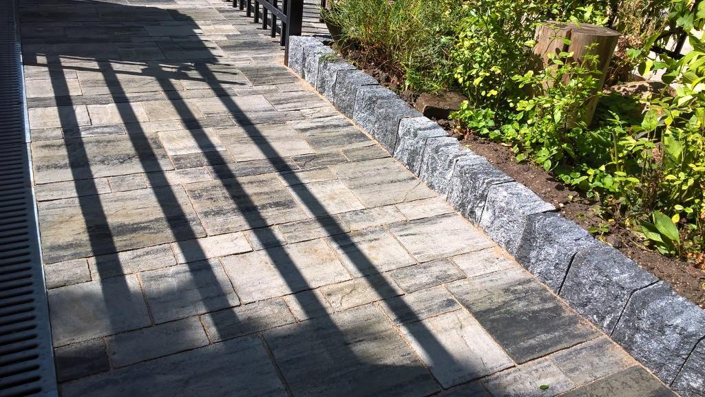 Ścieżka z kostki brukowej w odcieniach szarości, obramowana granitowymi kostkami, oświetlona promieniami słońca tworzącymi cienie, z widocznym odpływem liniowym.