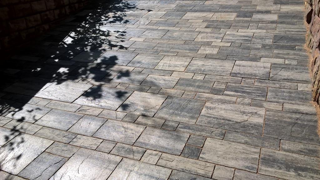 Ułożona szara kostka betonowa w różnych rozmiarach, z cieniem drzewa i fragmentem muru po bokach. Układ mozaikowy, widok z góry pod kątem.