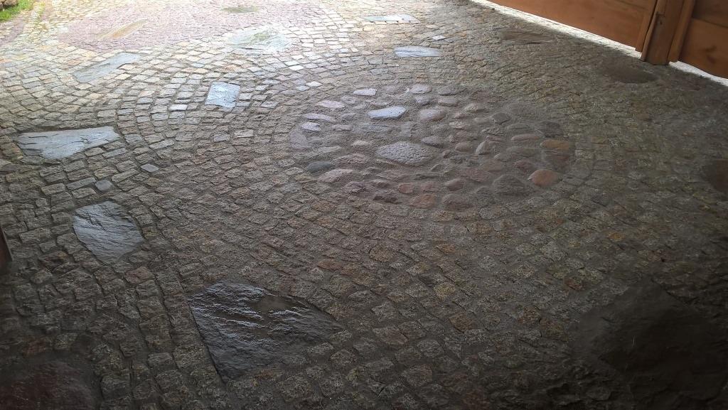 Ułożona, mokra nawierzchnia z małej kostki granitowej w odcieniach szarości i brązu, ułożona w łukowaty wzór. Fragment drewnianej konstrukcji w prawym górnym rogu.