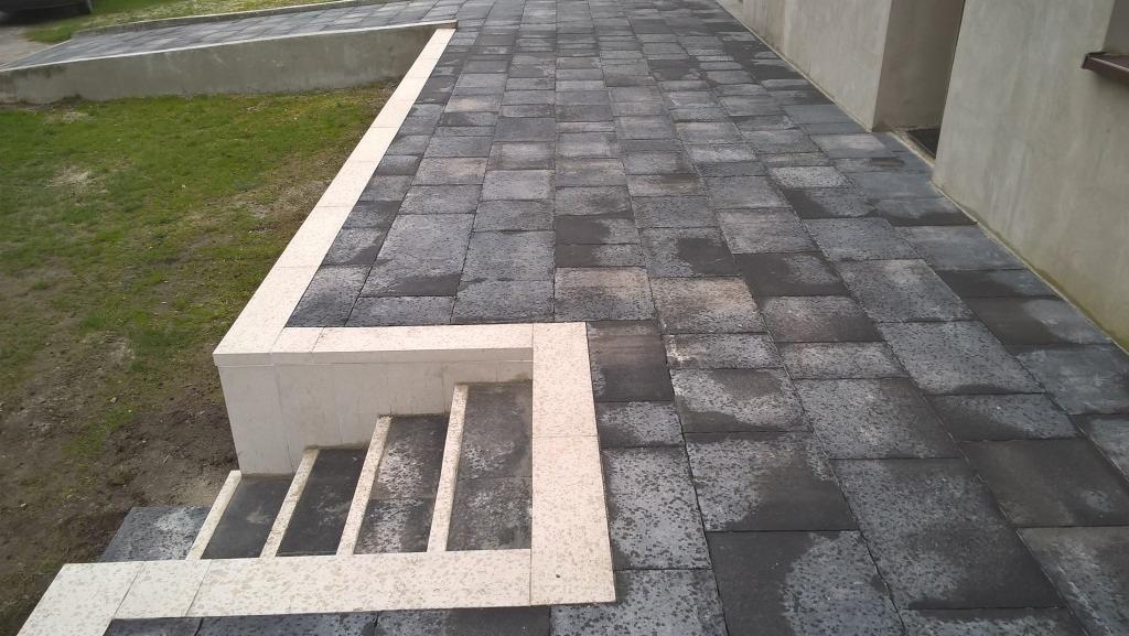 Ułożona, grafitowa nawierzchnia z kostki przy schodach i murku z jasnego kamienia. Widok z góry, podkreślający regularność wzoru i estetyczne wykończenie.