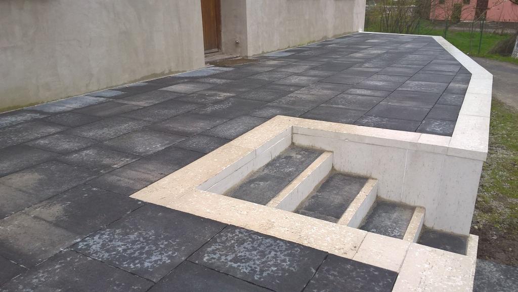 Ułożona, ciemna nawierzchnia z kostki przy budynku, z jasnymi schodami. Widoczne detale faktury kostki i wykończenia schodów.