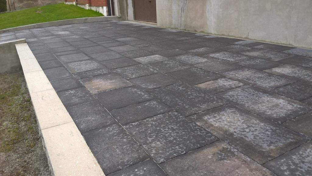 Czarno-szara kostka brukowa ułożona na podjeździe, z jasnym obrzeżem i ścianą budynku w tle. Widok na gotową nawierzchnię z kostki.