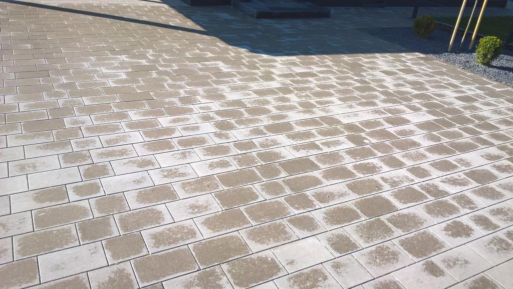 Ułożona, prostokątna kostka brukowa w odcieniach szarości i beżu, z widocznymi śladami wilgoci, tworząca wzór na podjeździe. W tle krzewy i elementy architektury.