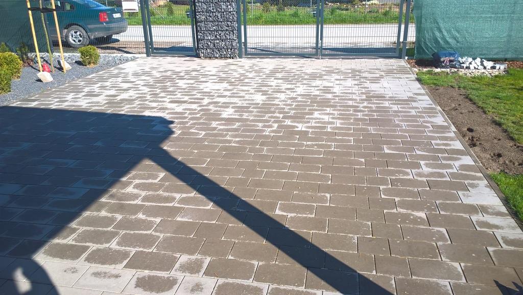 Ułożona kostka brukowa na podjeździe, widok z góry. Ciemnoszare prostokątne kostki, brama w tle. Częściowe zacienienie na powierzchni.