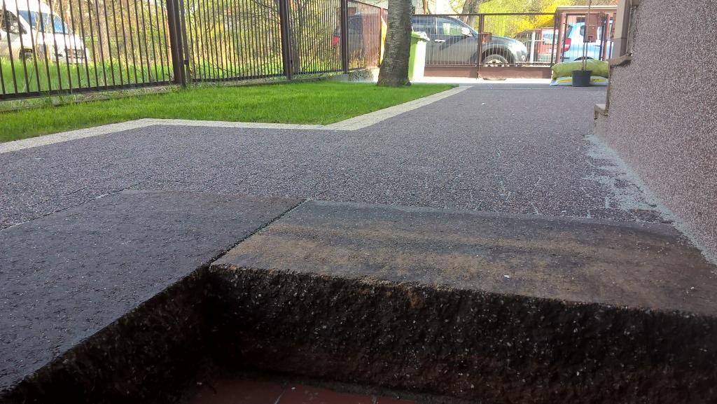 Nawierzchnia z żywicy epoksydowej z obrzeżem przy ogrodzeniu i budynku. Widoczne schody z kostki brukowej i trawnik. Szara kostka, brązowe ogrodzenie.