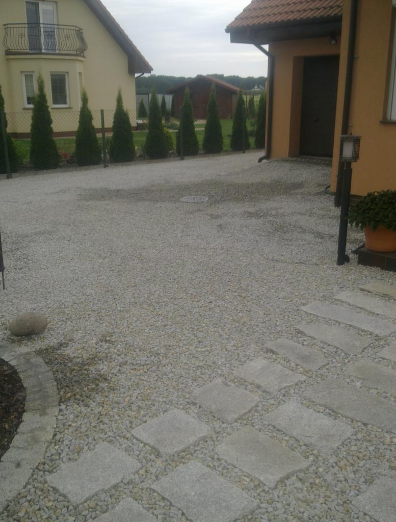 Żwirowa nawierzchnia podjazdu z elementami z płyt kamiennych i obrzeżem rabaty, tło: dom z balkonem i rząd iglaków.