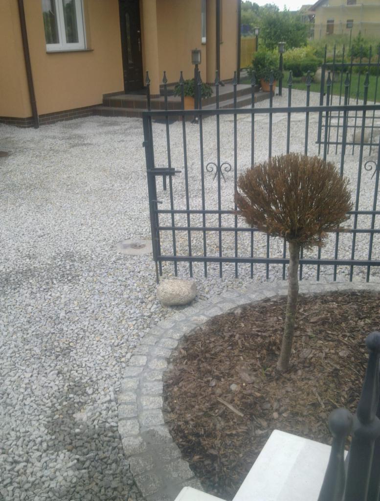 Żwirowa ścieżka przy domu, obrzeże z kostki granitowej okalające rabatę z drzewkiem, czarny metalowy płot. Ujęcie z niskiej perspektywy.