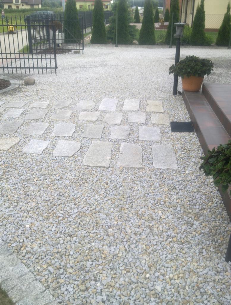 Ścieżka z nieregularnych płyt kamiennych ułożona na żwirowym podłożu. Widoczne obrzeża z kostki granitowej i elementy zieleni ogrodowej.