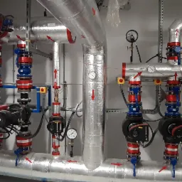 Złożona instalacja rur i pomp w srebrnej izolacji, z czerwonymi i niebieskimi elementami, widoczne manometry i strzałki wskazujące kierunek przepływu.