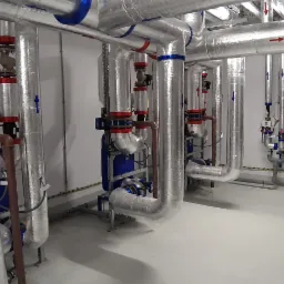 Rozbudowana instalacja rur w pomieszczeniu technicznym, rury izolowane folią aluminiową, liczne zawory i manometry w kolorach czerwonym i niebieskim, strzałki wskazujące kierunek przepływu.