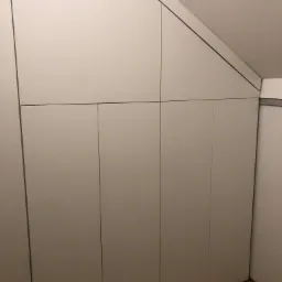 Biała szafa wnękowa z płaskimi drzwiami, dopasowana do skosu poddasza, na drewnianej podłodze.