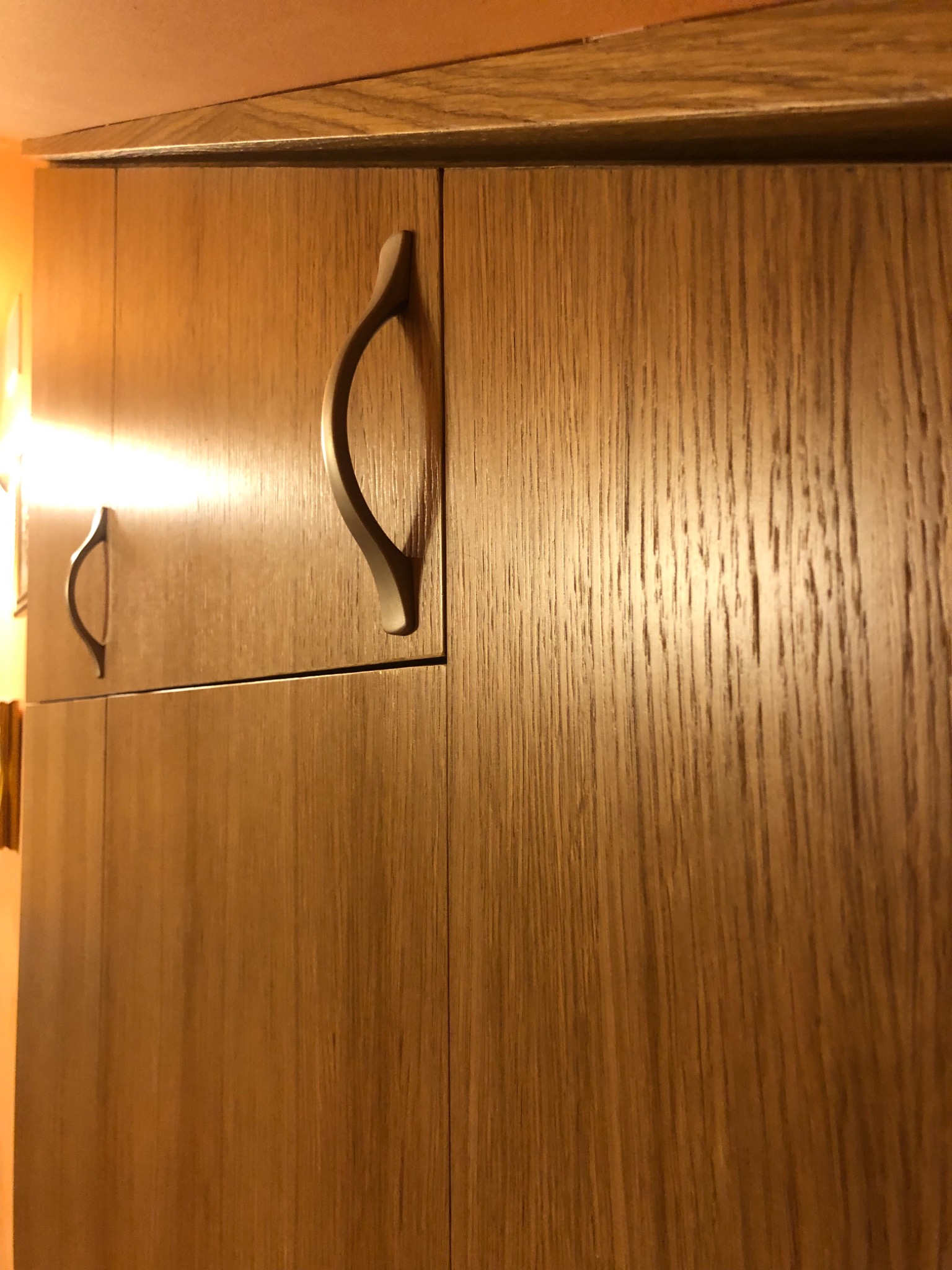 Fragment szafki kuchennej z drewnopodobnymi frontami i metalowymi uchwytami w kształcie łezki, oświetlonej ciepłym światłem.