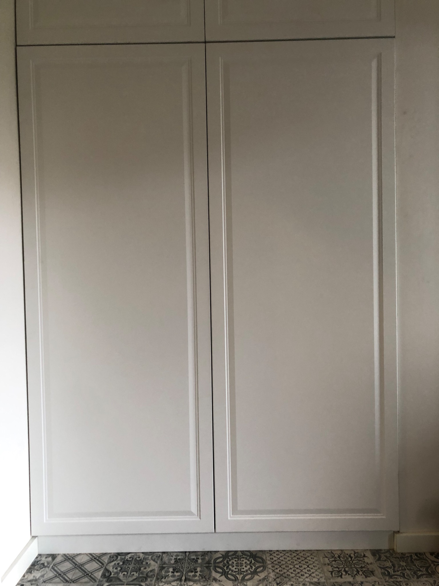 Białe, dwudrzwiowe szafy wnękowe z frezowanymi frontami, dolna krawędź widoczna nad wzorzystą podłogą.