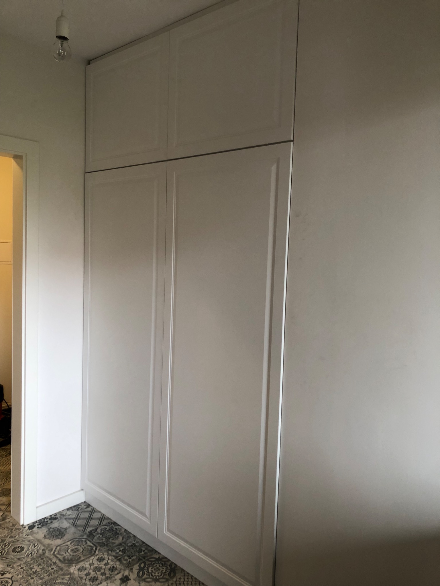 Biała, wysoka szafa z czterema drzwiami, z widocznym fragmentem podłogi z wzorzystymi płytkami oraz żarówką bez klosza na suficie.