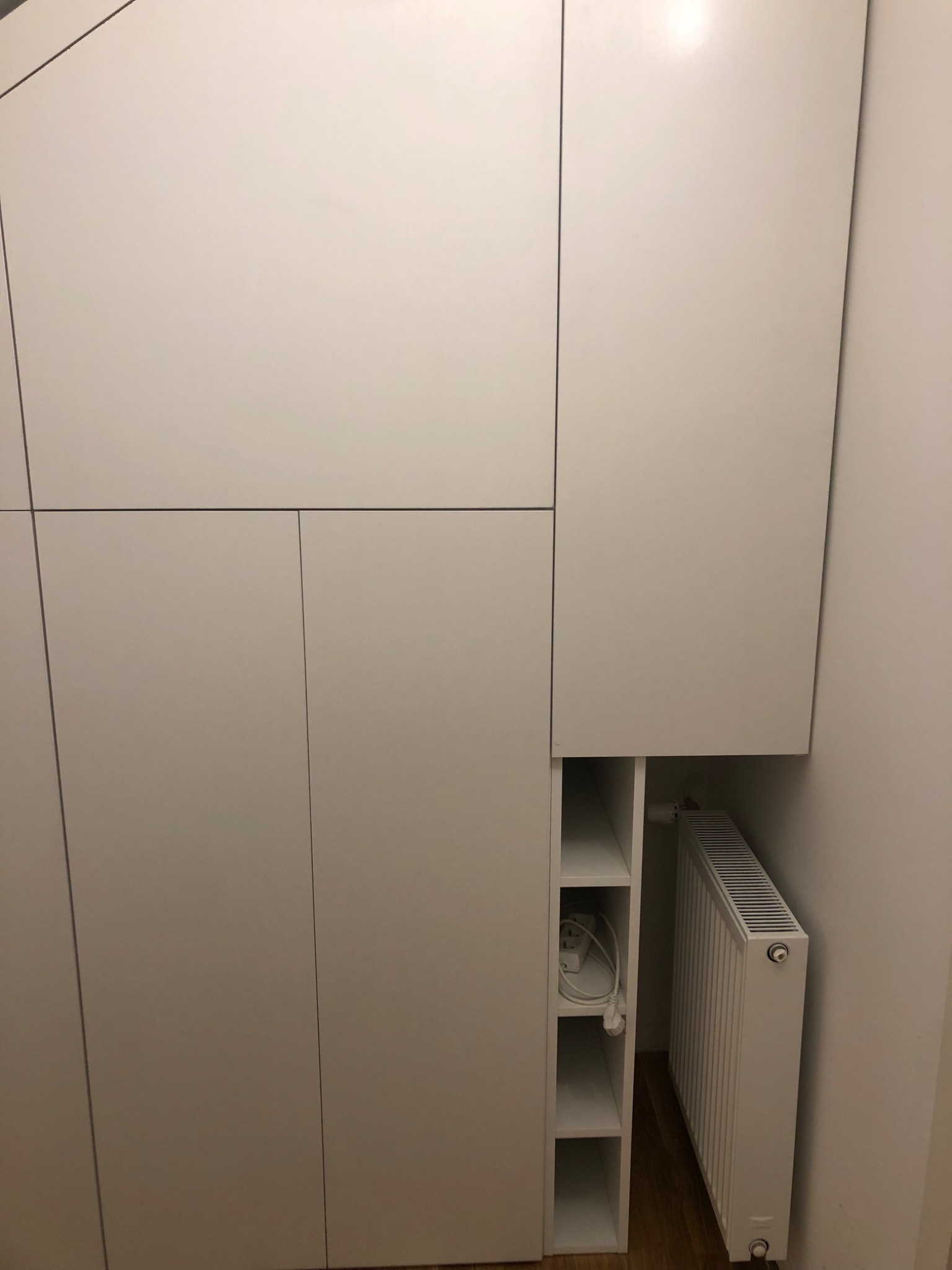 Biała szafa na wymiar z wnęką na grzejnik i półkami, widoczny przedłużacz w środku.