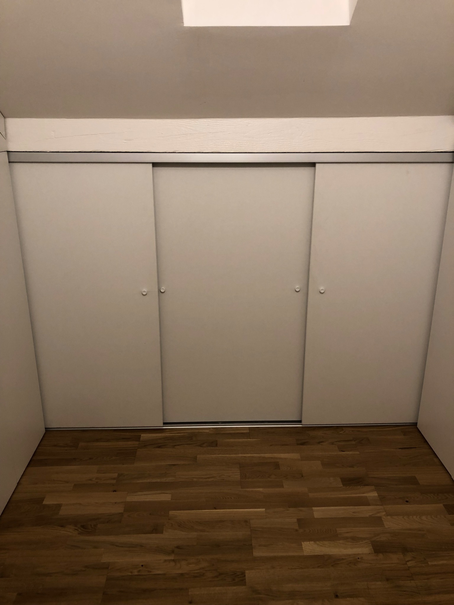 Wnętrze pokoju z drewnianą podłogą i trzydrzwiową szafą wnękową z przesuwnymi, białymi drzwiami.