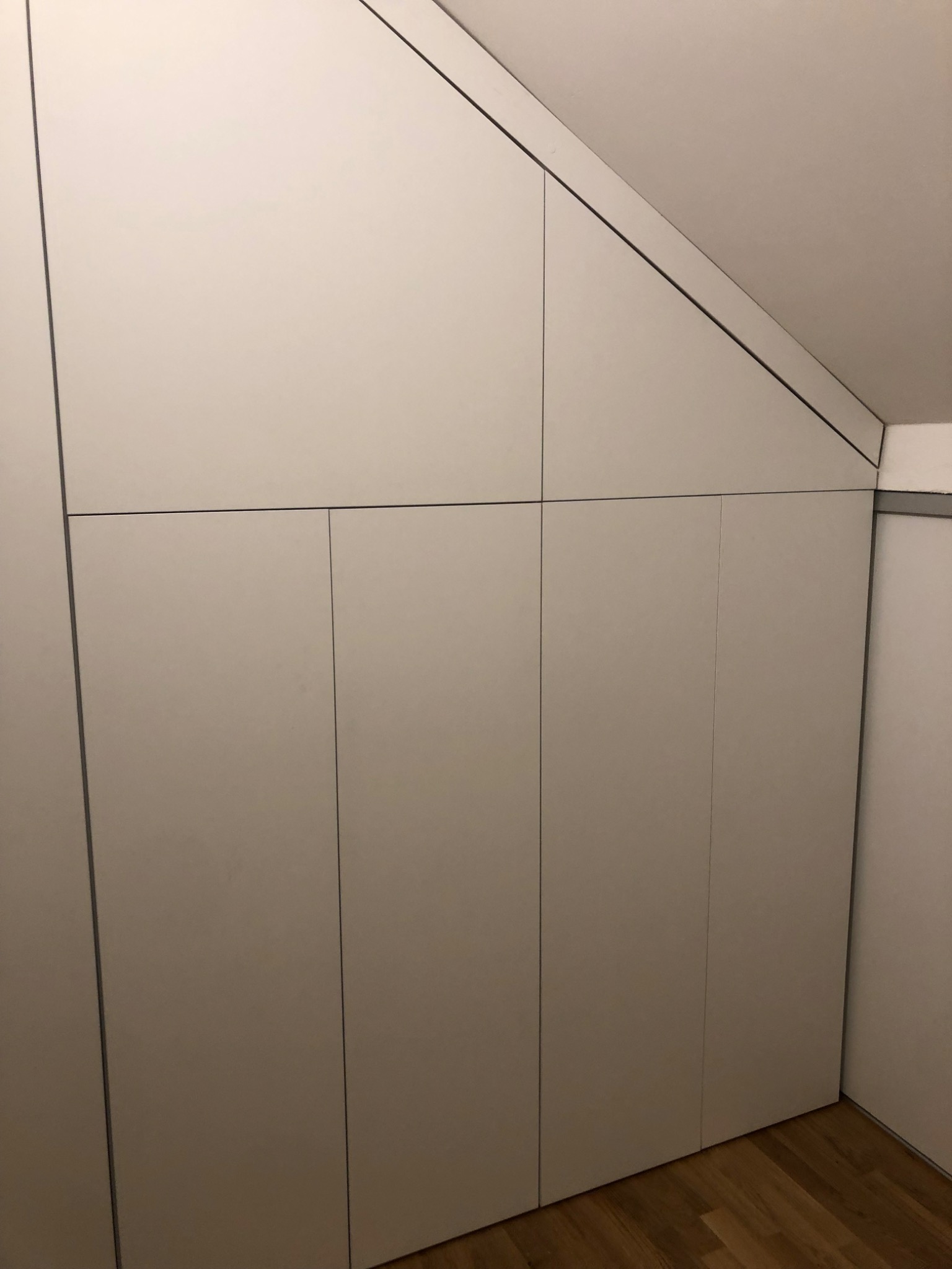 Biała szafa wnękowa z płaskimi drzwiami, dopasowana do skosu poddasza, na drewnianej podłodze.