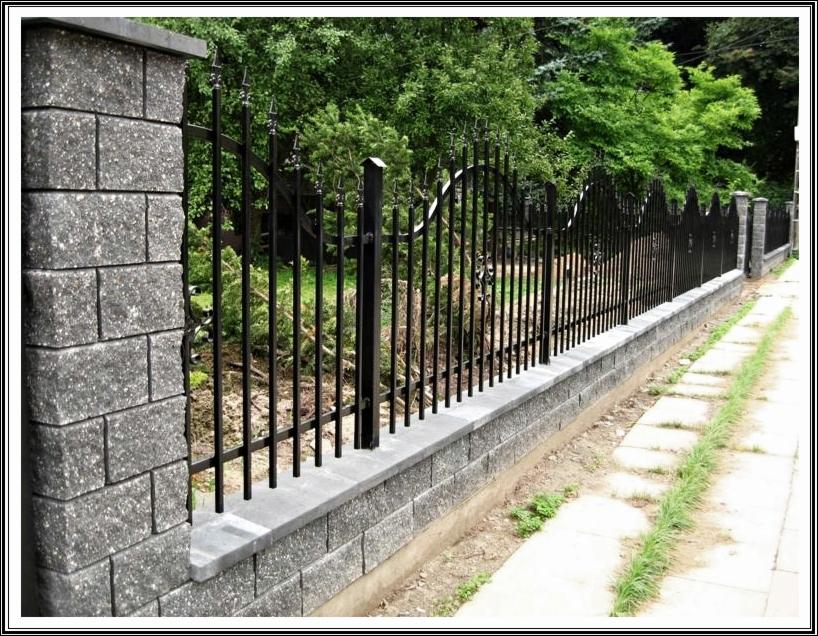 Eleganckie ogrodzenie z czarnych prętów i szarej, murowanej podmurówki, zwieńczone ozdobnymi elementami. Tło stanowi bujna zieleń drzew i krzewów.