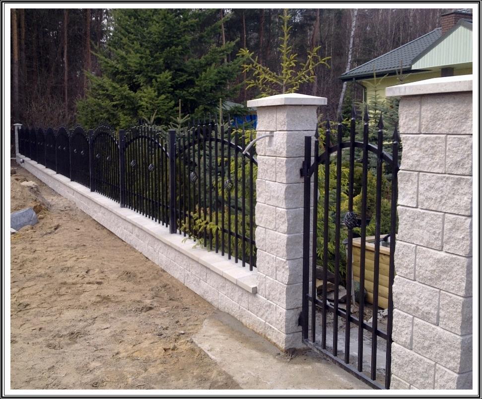 Eleganckie, czarne ogrodzenie z bramą, z kamiennymi słupkami i podmurówką, na tle zieleni. Charakterystyczne, dekoracyjne elementy w kształcie liści.
