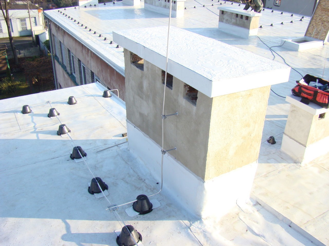 Biały dach płaski z zamontowaną instalacją odgromową, widoczne kominy z obróbkami blacharskimi i narzędzia dekarza