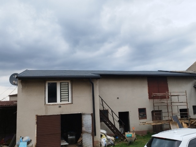 Szary dach z blachy na dwukondygnacyjnym budynku z beżową elewacją, widoczne rynny, antena satelitarna oraz rusztowania przy ścianie.