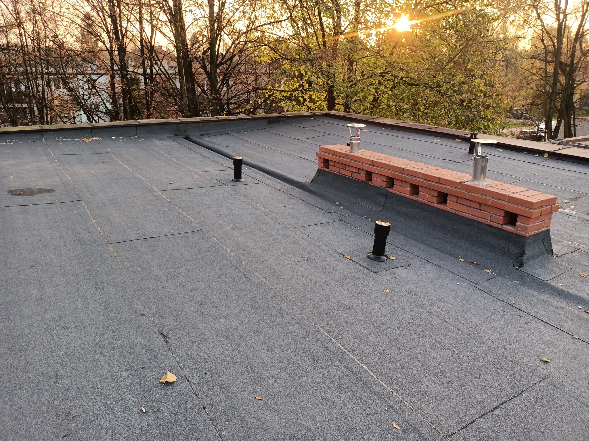 Płaski dach pokryty papą termozgrzewalną z wystającymi kominami wentylacyjnymi i kominem murowanym, widoczne jesienne liście na powierzchni, w tle drzewa w jesiennej scenerii podświetlone...