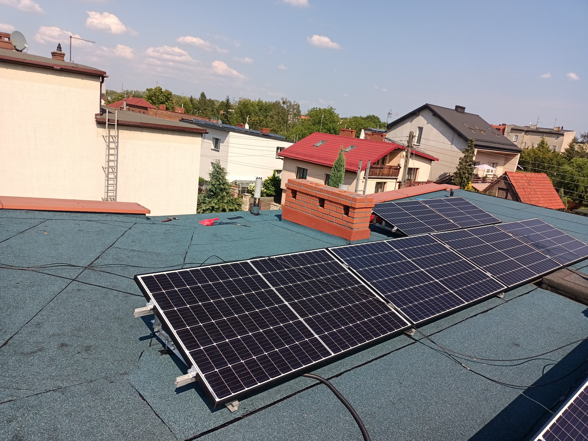 Panele słoneczne zamontowane na pokryciu dachowym z papy, widok z góry na tle zabudowy jednorodzinnej w słoneczny dzień.