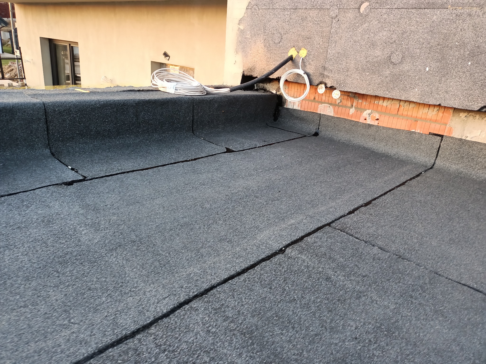 Czarny, płaski dach pokryty papą z widocznymi łączeniami, na dachu zwój białych kabli i czarna rura, w tle fragment budynku z oknem.