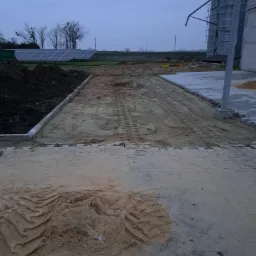 Przygotowanie terenu pod budowę: wyrównywanie piaskiem, widoczne ślady opon, w tle silosy zbożowe i panele słoneczne.