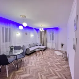 Nowoczesny salon z parkietem w jodełkę, szarym narożnikiem, szklanym stołem i nastrojowym niebieskim oświetleniem LED podsufitowym.