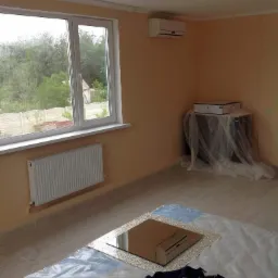 Pok&oacute;j przed malowaniem: zabezpieczone folią podłogi i meble, grzejnik, okno z widokiem na zieleń, klimatyzator na ścianie.