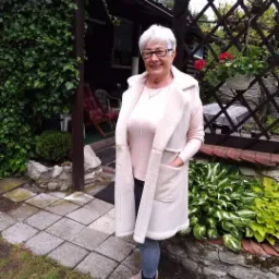 Uśmiechnięta kobieta w okularach, ubrana w długi, kremowy bezrękawnik z futerkiem, stoi na tle ogrodu z altaną porośniętą bluszczem i rabatą z funkiami.