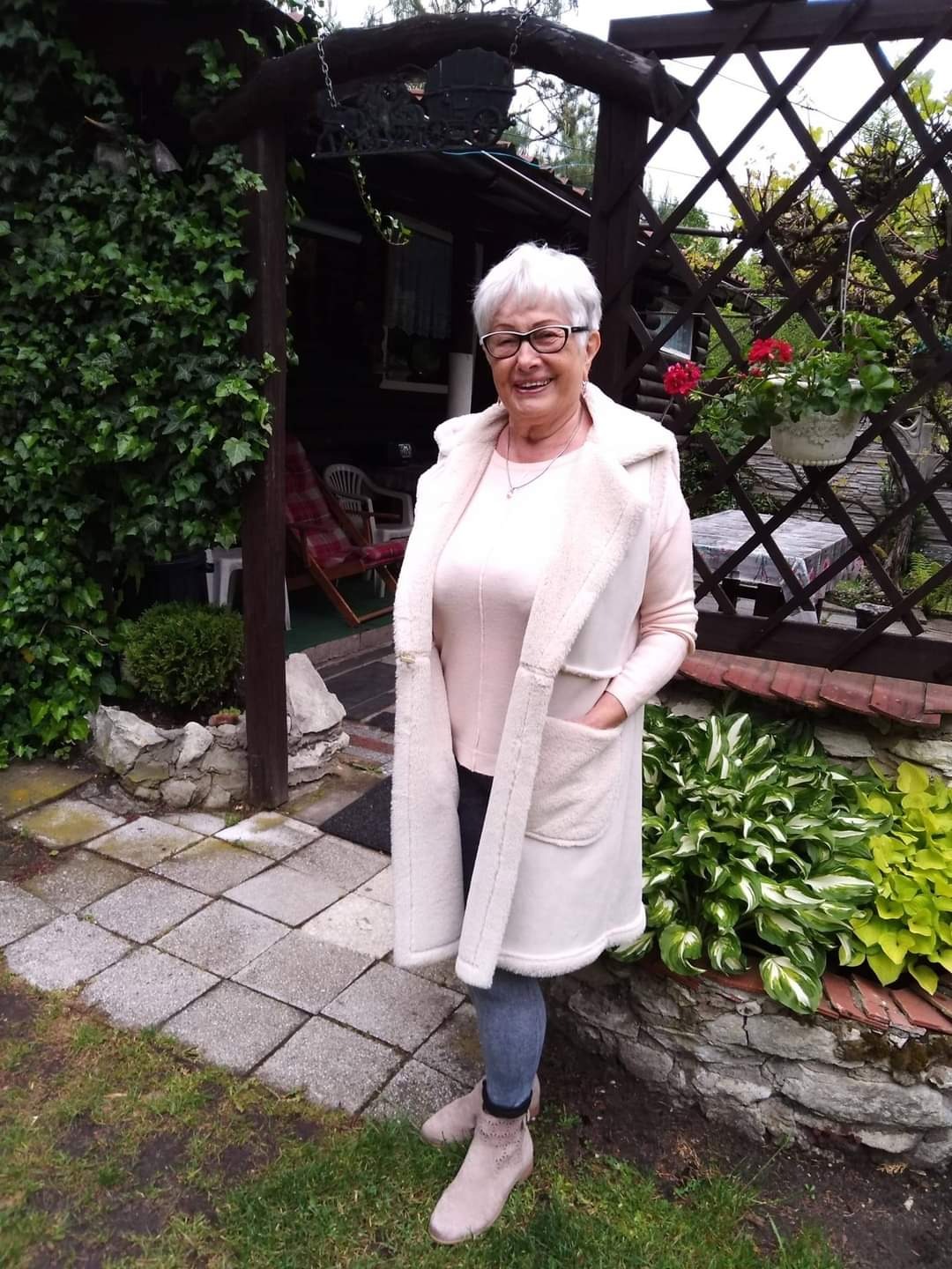 Uśmiechnięta kobieta w okularach, ubrana w długi, kremowy bezrękawnik z futerkiem, stoi na tle ogrodu z altaną porośniętą bluszczem i rabatą z funkiami.