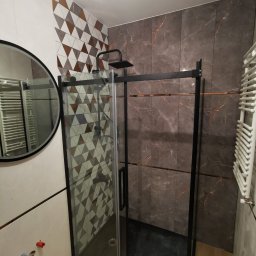 WOJBUD Firma Ogólnobudowlana - Nowoczesna kabina prysznicowa z czarną ramą i deszczownicą, obok okrągłe lustro, ściany wyłożone płytkami w geometryczne wzory i imitujące marmur z akcentami miedzianymi, grzejnik drabinkowy po...