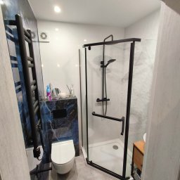 WOJBUD Firma Ogólnobudowlana - Nowoczesna łazienka z czarnymi akcentami: kabina prysznicowa z czarnymi ramami, grzejnik drabinkowy, wisząca toaleta i marmurowa ściana z czarną zabudową. Na blacie dekoracje i kosmetyki.