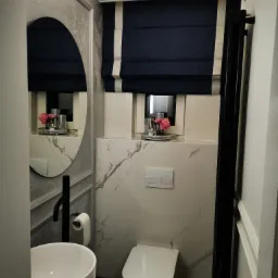 Nowoczesna łazienka z umywalką nablatową, toaletą wiszącą i grzejnikiem drabinkowym w czarnym kolorze, marmurowe płytki na ścianach i roleta rzymska w oknie