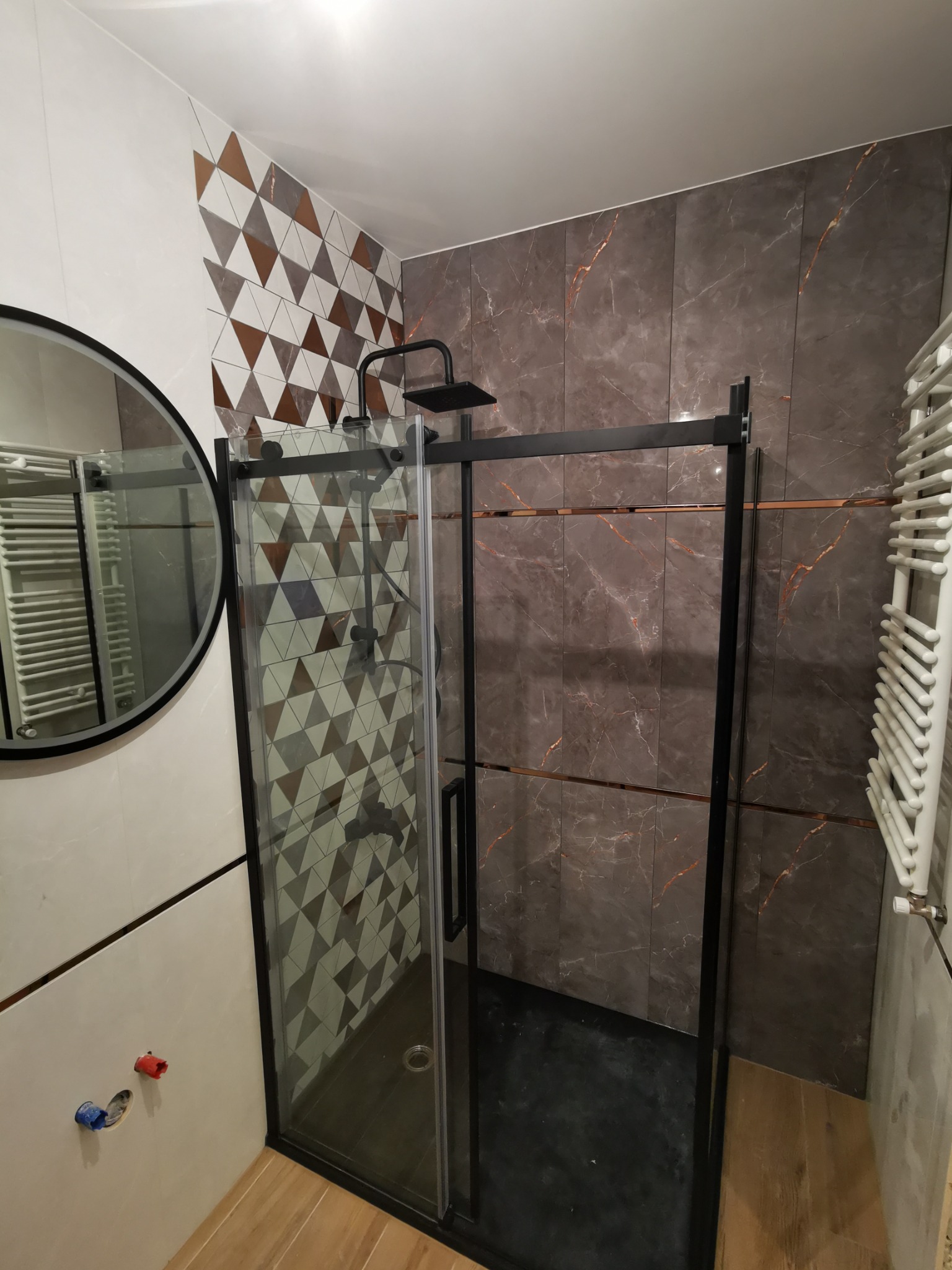 Nowoczesna kabina prysznicowa z czarną ramą i deszczownicą, obok okrągłe lustro, ściany wyłożone płytkami w geometryczne wzory i imitujące marmur z akcentami miedzianymi, grzejnik drabinkowy po...