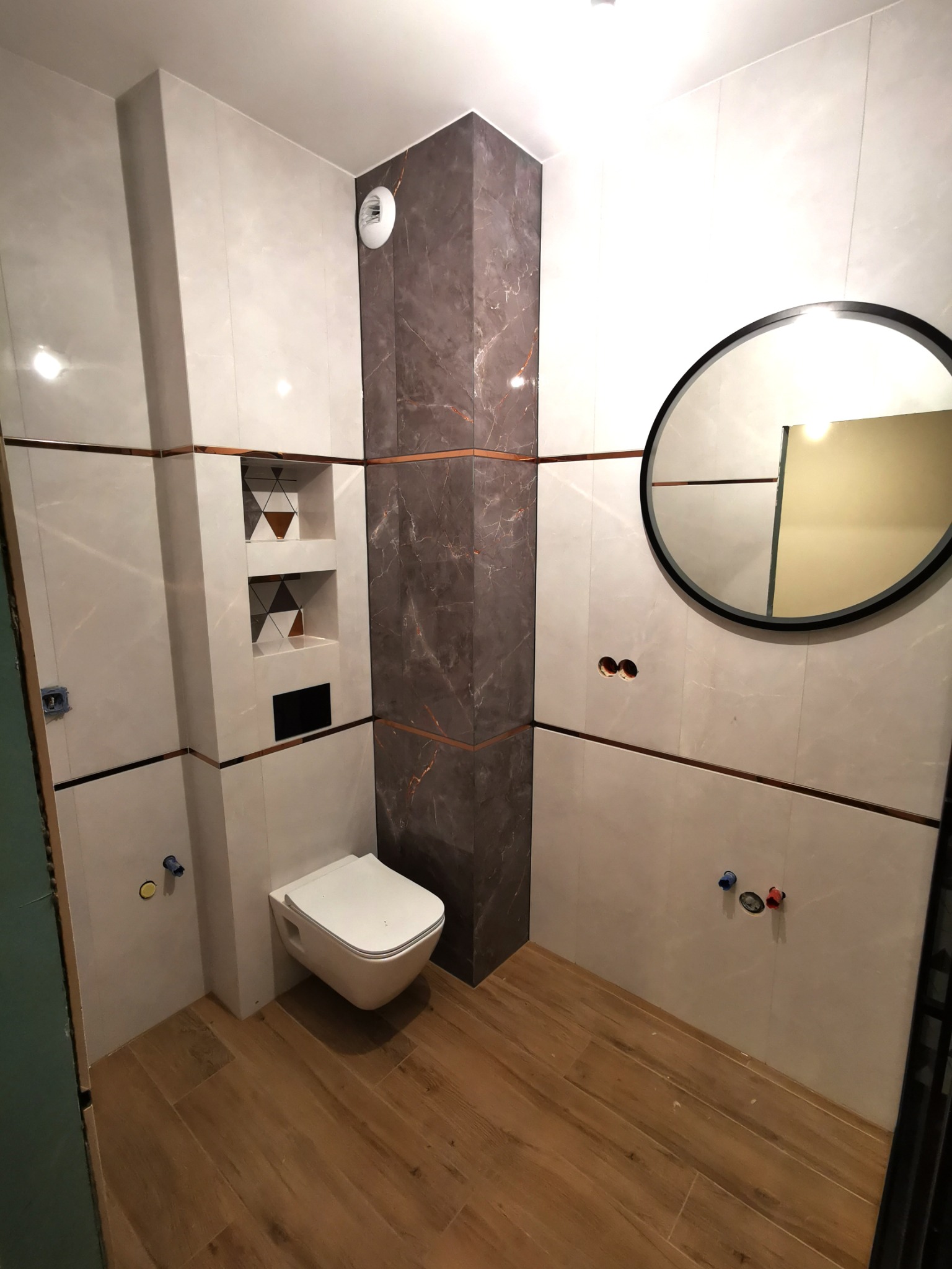 Nowoczesna łazienka z podwieszaną toaletą, marmurową kolumną i okrągłym lustrem, wykończona płytkami w odcieniach beżu i brązu z akcentami miedzianymi.