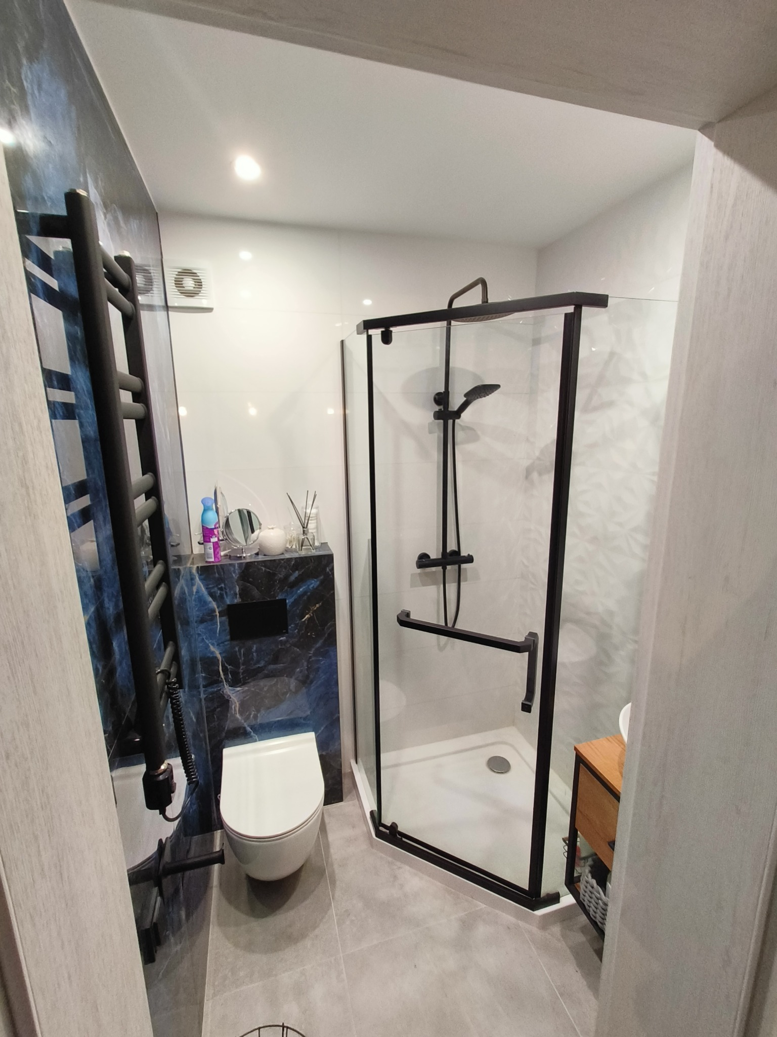 Nowoczesna łazienka z czarnymi akcentami: kabina prysznicowa z czarnymi ramami, grzejnik drabinkowy, wisząca toaleta i marmurowa ściana z czarną zabudową. Na blacie dekoracje i kosmetyki.