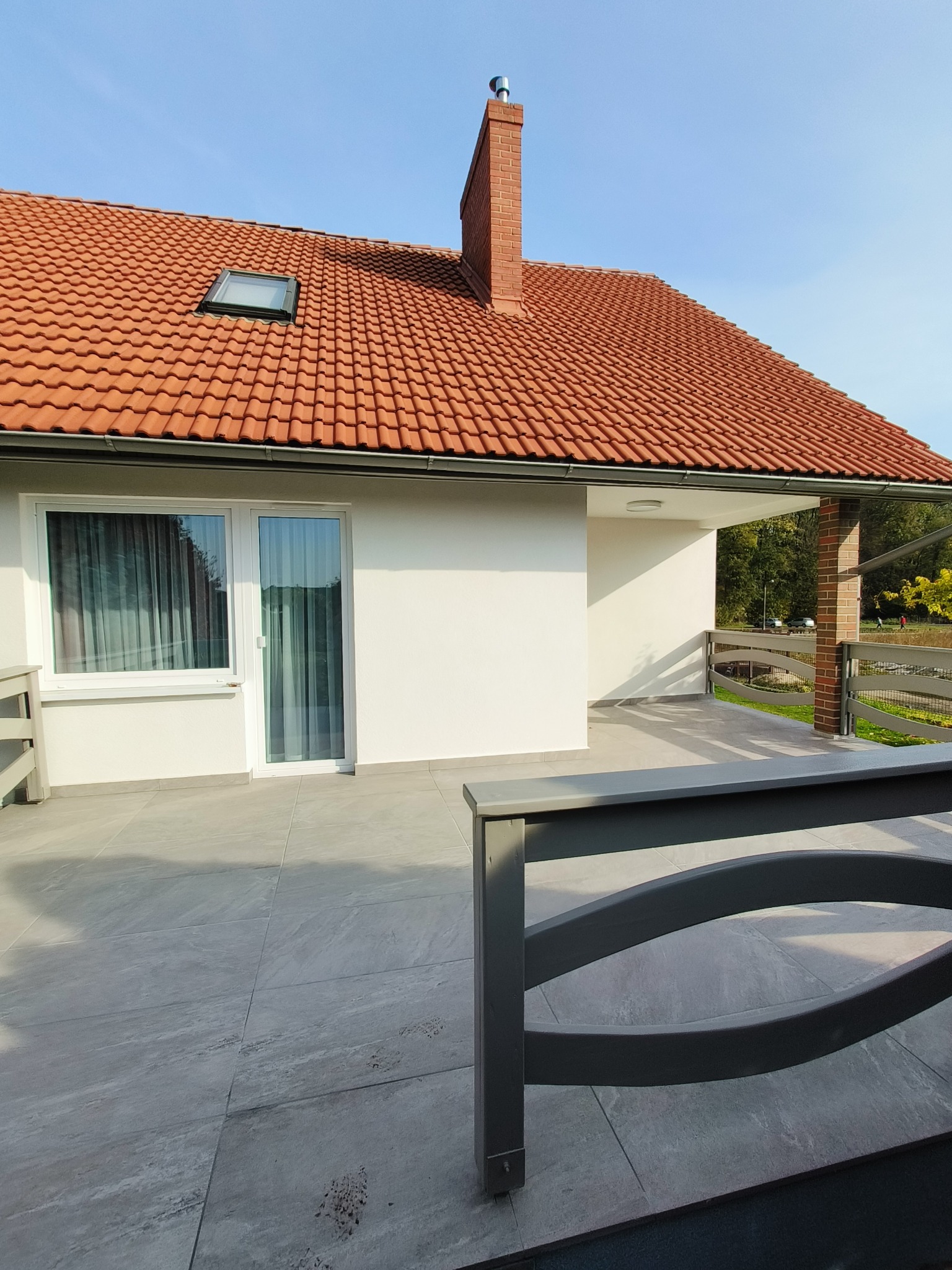 Nowoczesny dom jednorodzinny z tarasem wyłożonym szarą płytką, widok na elewację z oknem i drzwiami balkonowymi, czerwony dach z kominem i oknem dachowym, szara balustrada tarasu.