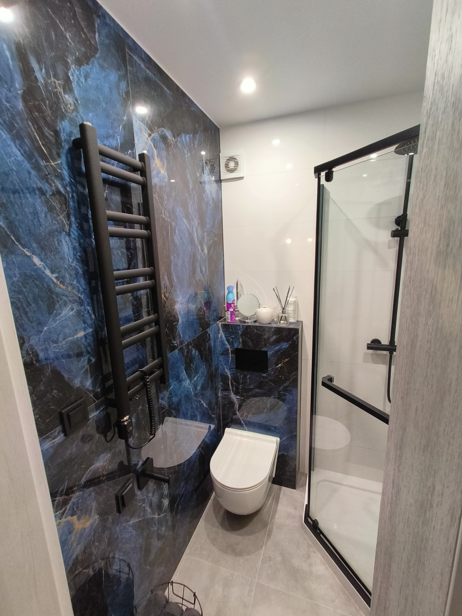 Nowoczesna łazienka z płytkami imitującymi marmur w odcieniach błękitu i złota, czarnym grzejnikiem drabinkowym i kabiną prysznicową z czarnymi profilami.