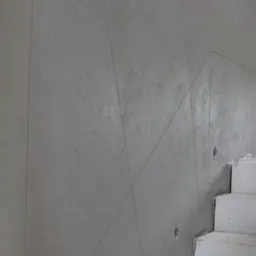 Surowa, szara ściana z tynkiem dekoracyjnym imitującym beton, widoczne pionowe i ukośne łączenia płyt oraz fragment betonowych schodów w minimalistycznym wnętrzu.
