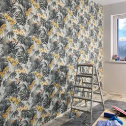 Pokój w trakcie dekorowania tapetą w tropikalne liście i żółte kwiaty, drabina aluminiowa stoi przy ścianie, podłoga zabezpieczona folią malarską, widoczne narzędzia i torba z akcesoriami.