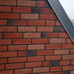 Fragment elewacji budynku z cegły klinkierowej w odcieniach czerwieni i brązu, z widoczną szarą listwą okapową i fragmentem rusztowania w tle.