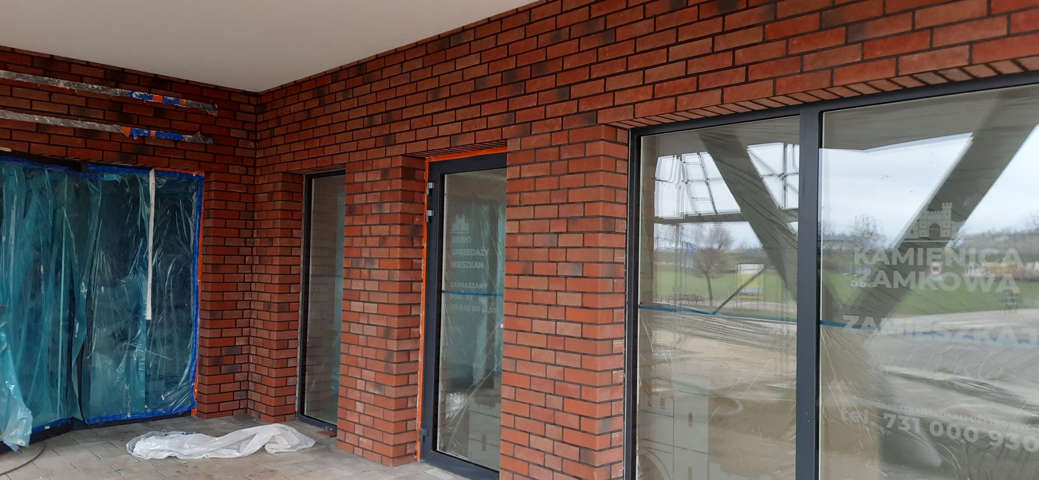 Murowana elewacja budynku z cegły klinkierowej w odcieniach czerwieni i brązu, widoczne okna i drzwi zabezpieczone folią ochronną, częściowo zakryte plandeką. Na szybie okna widoczny napis...