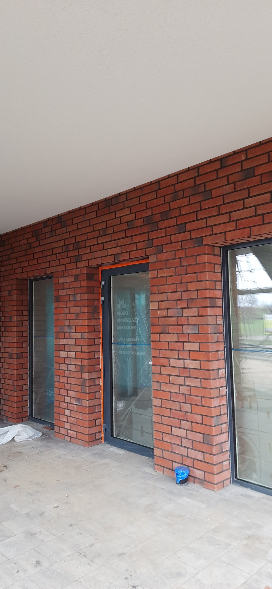 Nowa elewacja budynku wykończona czerwoną cegłą klinkierową, z zamontowanymi oknami i drzwiami zabezpieczonymi folią, widoczne ślady prac wykończeniowych na chodniku.