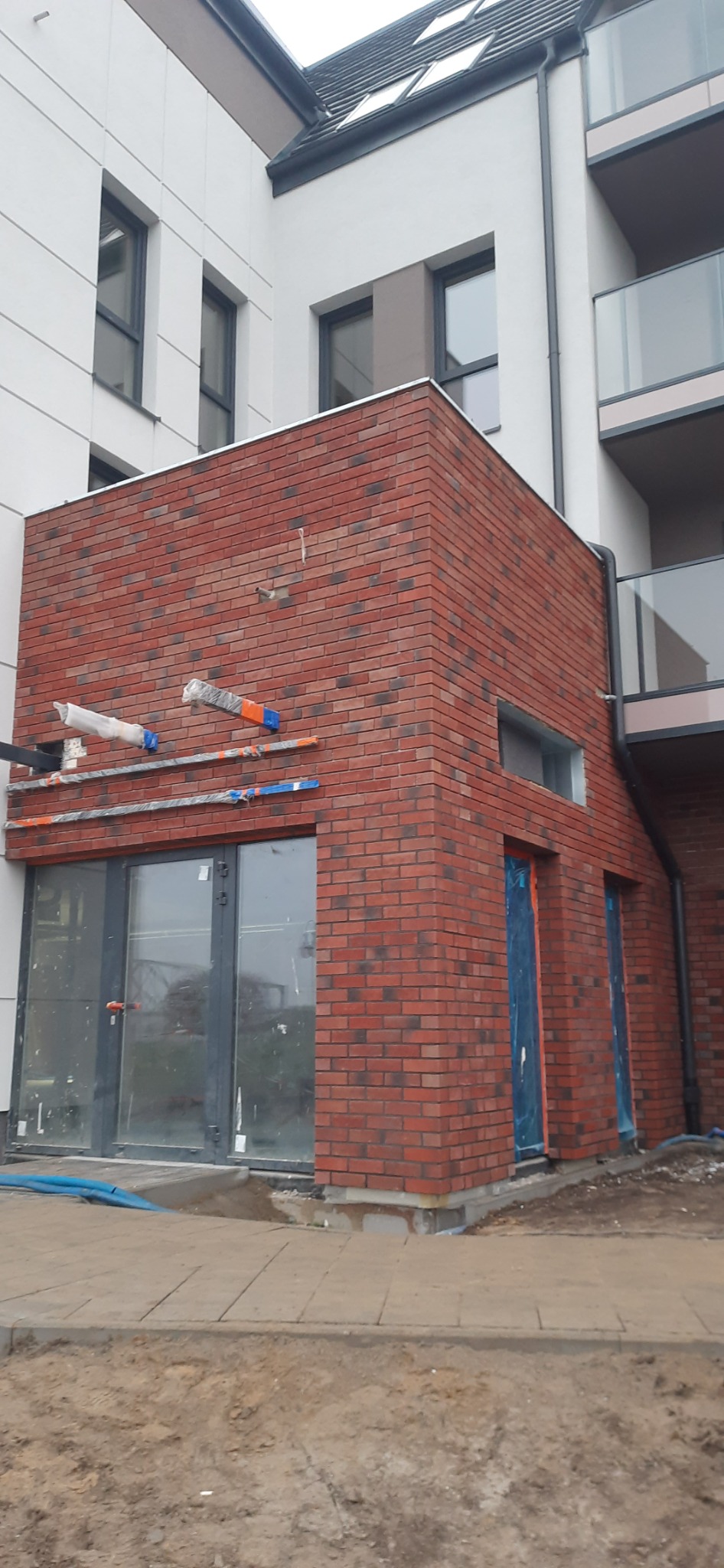 Ceglana elewacja budynku w trakcie budowy z widocznymi oknami i drzwiami zabezpieczonymi folią, oraz elementami rusztowania.