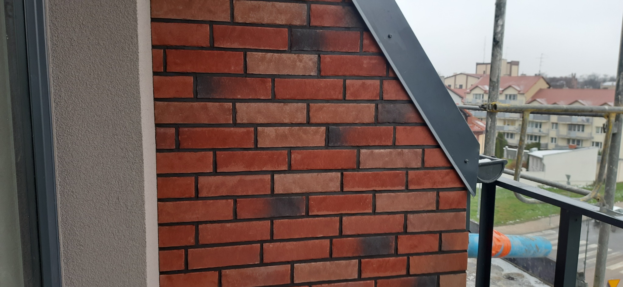 Fragment elewacji budynku z cegły klinkierowej w odcieniach czerwieni i brązu, z widoczną szarą listwą okapową i fragmentem rusztowania w tle.